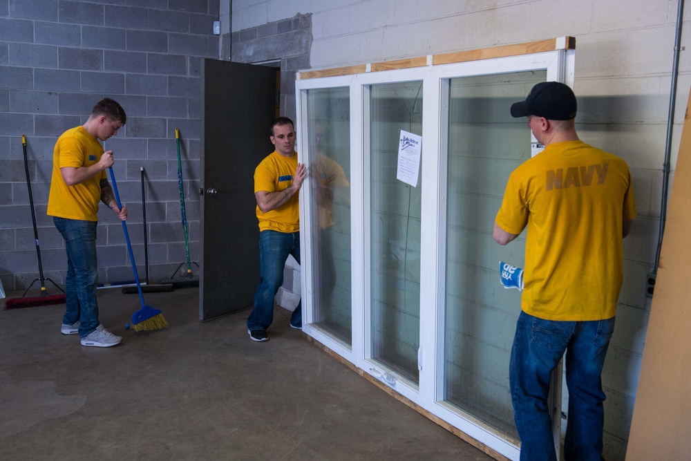Three men wearing matching yellow t-shirts are working inside a concrete block building. Two of them are handling a large white-framed sliding glass window/door unit, while the third is sweeping the floor with a broom. The man on the right has 'NAVY' printed on the back of his shirt, suggesting these may be Navy personnel performing volunteer or community service work.