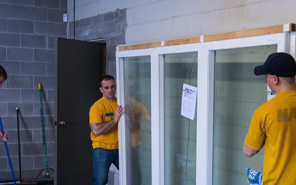 Three men wearing matching yellow t-shirts are working inside a concrete block building. Two of them are handling a large white-framed sliding glass window/door unit, while the third is sweeping the floor with a broom. The man on the right has 'NAVY' printed on the back of his shirt, suggesting these may be Navy personnel performing volunteer or community service work.