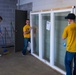Three men wearing matching yellow t-shirts are working inside a concrete block building. Two of them are handling a large white-framed sliding glass window/door unit, while the third is sweeping the floor with a broom. The man on the right has 'NAVY' printed on the back of his shirt, suggesting these may be Navy personnel performing volunteer or community service work.