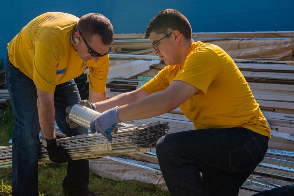 Two individuals wearing matching yellow t-shirts are working together outdoors, handling metal construction materials or ceiling grid components. They appear to be volunteers or workers engaged in a construction or cleanup project, with one person wearing sunglasses and gloves. A pile of building materials, including what appears to be ceiling tiles and framing, is stacked in the background.