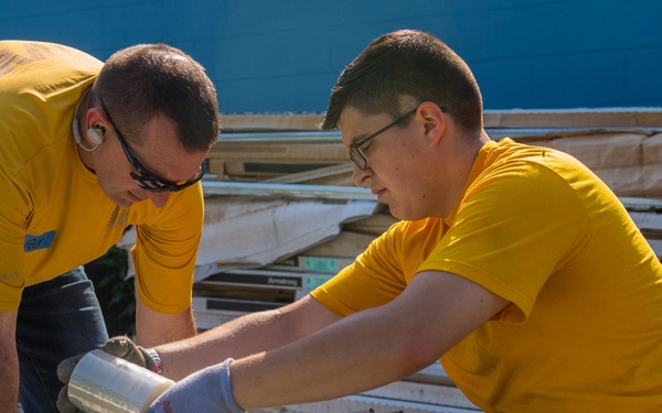 Two individuals wearing matching yellow t-shirts are working together outdoors, handling metal construction materials or ceiling grid components. They appear to be volunteers or workers engaged in a construction or cleanup project, with one person wearing sunglasses and gloves. A pile of building materials, including what appears to be ceiling tiles and framing, is stacked in the background.