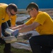 Two individuals wearing matching yellow t-shirts are working together outdoors, handling metal construction materials or ceiling grid components. They appear to be volunteers or workers engaged in a construction or cleanup project, with one person wearing sunglasses and gloves. A pile of building materials, including what appears to be ceiling tiles and framing, is stacked in the background.
