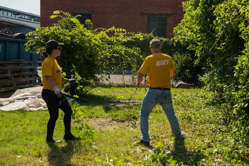 Two individuals wearing matching yellow shirts are performing outdoor cleanup or landscaping work in an overgrown urban lot. The person on the right has 'NAVY' printed on the back of their shirt, suggesting this may be a military community service or volunteer event. They are clearing brush and branches near a red brick building.