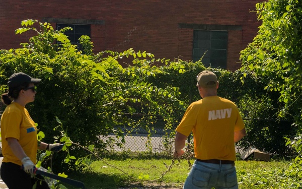 Two individuals wearing matching yellow shirts are performing outdoor cleanup or landscaping work in an overgrown urban lot. The person on the right has 'NAVY' printed on the back of their shirt, suggesting this may be a military community service or volunteer event. They are clearing brush and branches near a red brick building.