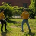 Two individuals wearing matching yellow shirts are performing outdoor cleanup or landscaping work in an overgrown urban lot. The person on the right has 'NAVY' printed on the back of their shirt, suggesting this may be a military community service or volunteer event. They are clearing brush and branches near a red brick building.