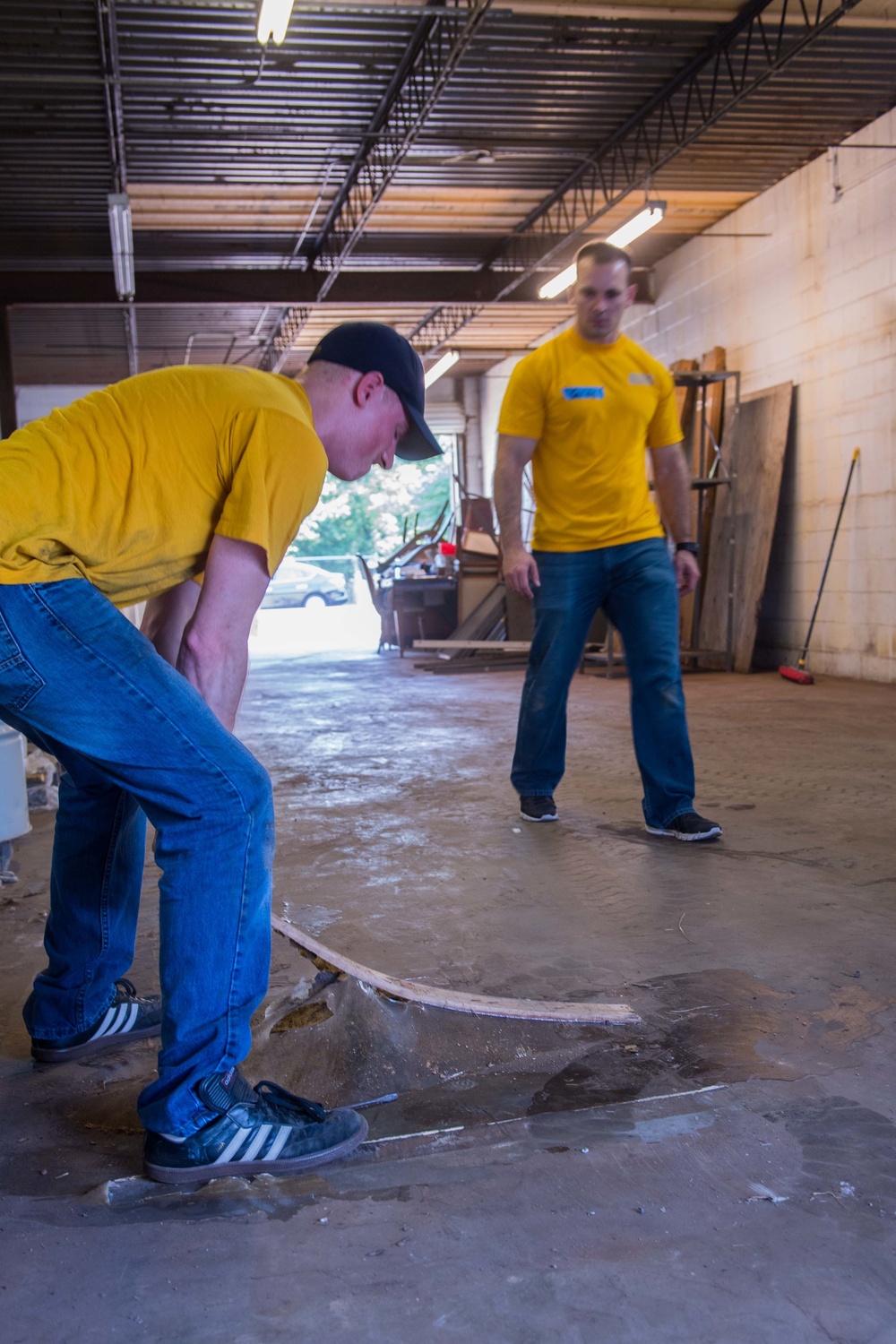 Two men wearing matching yellow t-shirts and blue jeans are working inside what appears to be a warehouse or industrial space. The foreground worker is bent over prying up or removing a piece of plywood or flooring material from a concrete floor. The background worker is standing upright, observing or supervising the task.