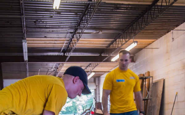 Two men wearing matching yellow t-shirts and blue jeans are working inside what appears to be a warehouse or industrial space. The foreground worker is bent over prying up or removing a piece of plywood or flooring material from a concrete floor. The background worker is standing upright, observing or supervising the task.