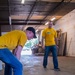 Two men wearing matching yellow t-shirts and blue jeans are working inside what appears to be a warehouse or industrial space. The foreground worker is bent over prying up or removing a piece of plywood or flooring material from a concrete floor. The background worker is standing upright, observing or supervising the task.