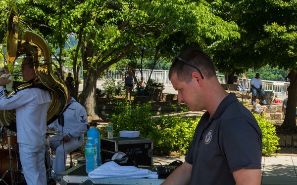 A man in a dark polo shirt and khaki shorts operates a Yamaha audio mixing console mounted in a road case labeled 'Government Property' at an outdoor event. In the background, a musician in a white U.S. Navy uniform plays a tuba, suggesting this is a U.S. Navy Fleet Band performance. The event appears to be taking place in a park or waterfront area on a sunny day.