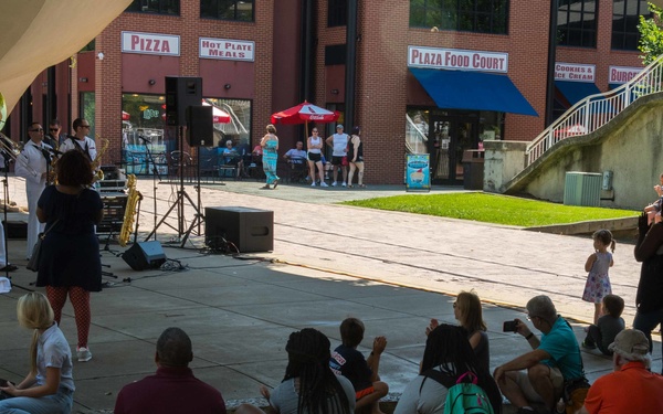 An outdoor public concert is taking place at a plaza food court area, where a military band dressed in white uniforms is performing for a crowd of spectators. Audience members are seated on the ground or standing around a paved plaza area, watching the performance. The setting appears to be a commercial outdoor venue with food court signage visible in the background.
