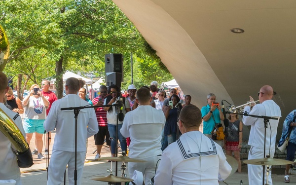 A military band, likely U.S. Navy musicians dressed in white uniforms, performs an outdoor concert at what appears to be a public park or plaza. The ensemble includes brass instruments, drums, and a tuba, playing beneath a curved architectural canopy structure. A diverse crowd of spectators watches and records the performance on their phones.