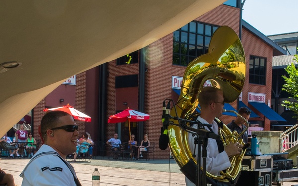 A U.S. Navy band performs an outdoor concert in what appears to be a public plaza or waterfront area. A sailor in white dress uniform plays a drum kit in the foreground, while another musician plays a sousaphone/tuba in the background. The setting includes brick buildings, outdoor dining umbrellas, and a small audience.