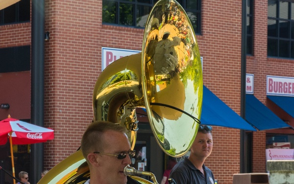 A U.S. Navy musician in white dress uniform plays a sousaphone (tuba) at an outdoor public performance. The large brass instrument wraps around the performer, who is wearing sunglasses and military ribbons on his chest. The setting appears to be a urban plaza or street festival near commercial buildings.