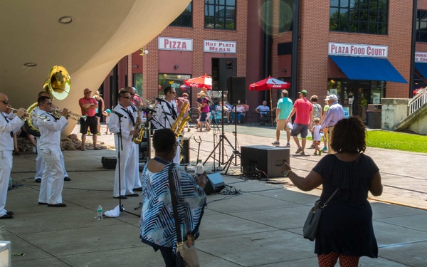 An outdoor concert is taking place at a public plaza amphitheater, where a military band dressed in white uniforms performs brass and wind instruments on a stage. Audience members are gathered around, with some standing and dancing near the performance area while others sit on stone steps. The background shows a brick building housing a Plaza Food Court with signs for Pizza, Burgers, and Hot Plate Meals.