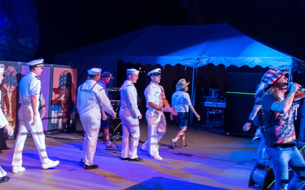 An outdoor nighttime concert or performance event featuring several individuals in white U.S. Navy dress uniforms walking across a stage alongside a flamboyantly dressed performer who appears to be a rock musician. The stage is illuminated with blue and purple lighting, with speaker stacks, a drum kit, and a large white tent visible in the background. The scene suggests a military appreciation or patriotic-themed entertainment event.