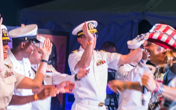 A group of U.S. Navy personnel in white dress uniforms are dancing and clapping at what appears to be an outdoor public event or festival. They are interacting with civilians, including a person wearing a red and white striped patriotic hat. The atmosphere is festive and celebratory, likely a community or military appreciation event.