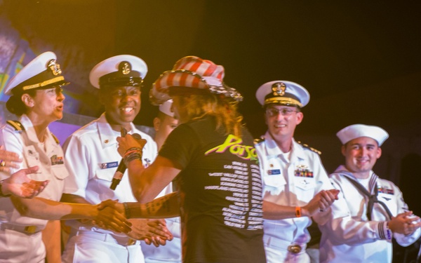 A performer wearing a patriotic top hat and a black 'Poison' tour shirt is on stage shaking hands with U.S. Navy officers and enlisted sailors in dress white uniforms. The scene appears to be a military appreciation concert or USO-style event, with the performer engaging directly with service members on stage. A civilian in a blue polo shirt with a lanyard is also present on the left side of the stage.