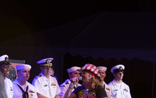 A performer wearing a patriotic red, white, and blue top hat and a graphic t-shirt is singing into a microphone on a stage, surrounded by several U.S. Navy personnel in white dress uniforms. The event appears to be a military appreciation or patriotic concert held under a tent. The performer is energetically engaging with the audience while Navy sailors stand behind him on stage.