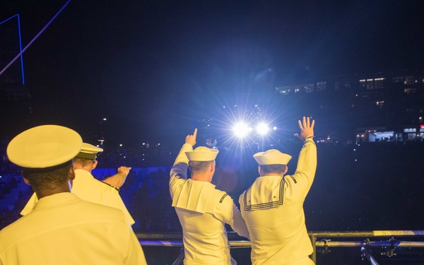 A group of U.S. Navy personnel in white dress uniforms are standing on what appears to be a stage or elevated platform at a large nighttime event, waving and cheering toward a crowd in a stadium or arena. The scene is brightly lit with stage lighting against a dark background, with a large audience visible below. A small American flag is visible among the crowd in the lower left area.