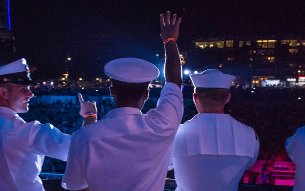 Four individuals wearing white U.S. Navy uniforms and white sailor caps are standing at a railing overlooking a large nighttime outdoor event or concert venue. They are facing away from the camera, with at least two of them raising their hands toward the crowd below. The scene is illuminated by colorful stage lighting in blue and pink/purple tones.