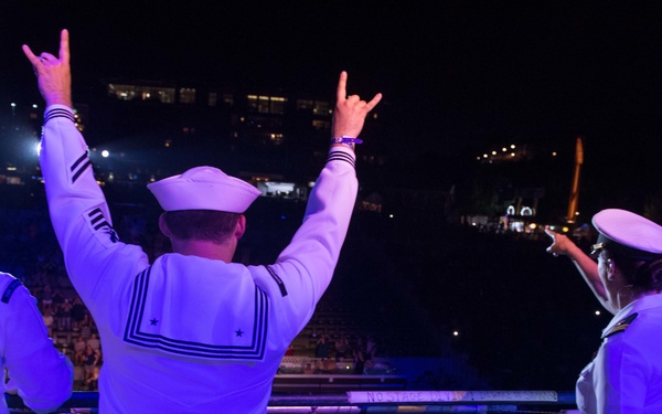 Four U.S. Navy sailors in white dress uniforms are standing on an elevated platform or balcony overlooking a large outdoor crowd at a nighttime event. Two sailors in the center have their arms raised, making 'rock on' or 'hook 'em horns' hand gestures toward the crowd below. The scene is illuminated by purple/blue stage lighting, suggesting a concert or public entertainment event.