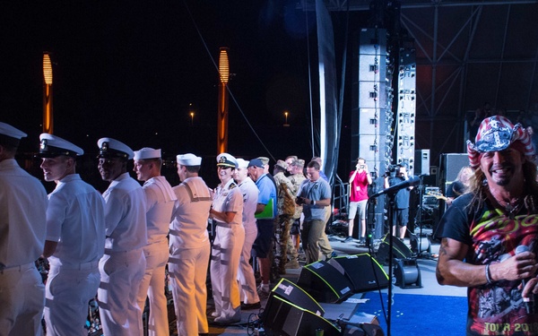 A concert stage scene at night featuring a rock performer wearing a decorated cap and graphic t-shirt standing at the front right of the stage. A line of military personnel in white Navy dress uniforms are assembled on the left side of the stage, suggesting a military appreciation or tribute moment during the performance. A large outdoor amphitheater audience is visible in the background.