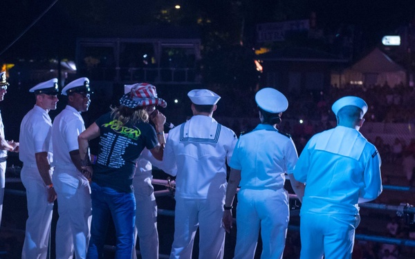 A nighttime outdoor concert or event stage featuring several U.S. Navy personnel in white dress uniforms alongside a civilian performer wearing a black band t-shirt and an American flag-themed hat. Stage monitor speakers with neon green trim are visible in the foreground, with a large crowd visible in the background. The setting appears to be a patriotic or military appreciation concert event.