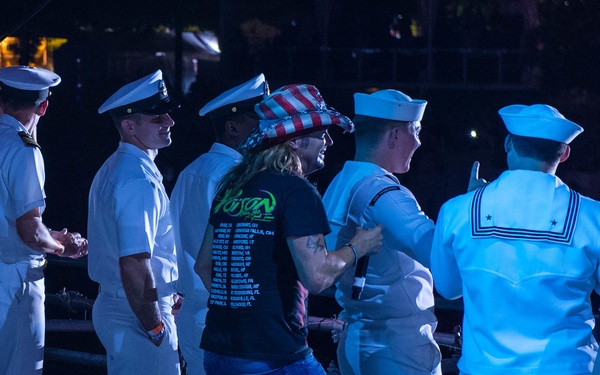 A group of U.S. Navy personnel in white dress uniforms are gathered at what appears to be an outdoor concert or military event at night. Among them stands a civilian wearing a black 'Poison' band t-shirt and an American flag-style top hat, appearing to interact enthusiastically with the sailors. Stage lighting illuminates the scene with a blue tint, and a 'POISON' concert sign is partially visible in the lower right corner.