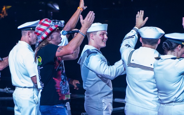 A group of U.S. Navy personnel in white dress uniforms are on stage at what appears to be a concert or entertainment event. A civilian performer wearing jeans, a black t-shirt, and an American flag-themed hat is interacting with the sailors, with several people raising their hands. A senior officer in a white service dress uniform with gold insignia stands to the left observing the scene.