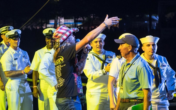 A group of U.S. Navy personnel in white uniforms are gathered on what appears to be a concert stage alongside civilians, including a performer wearing a dark t-shirt and an American flag bandana with arm raised. Stage monitors and audio equipment are visible in the foreground, suggesting this is a live music or entertainment event. The setting appears to be an outdoor nighttime venue, likely a military appreciation concert or Fleet Week-type event.