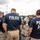 A large group of law enforcement officers are gathered in an outdoor area, likely a parking lot or staging area. Multiple officers are wearing navy blue t-shirts identifying them as 'POLICE HSI' (Homeland Security Investigations) and 'POLICE - Border Enforcement Security Taskforce (BEST)'. The gathering appears to be a pre-operation briefing or assembly, with many officers facing toward a central point.