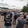 A large gathering of law enforcement personnel in an outdoor parking lot area, with multiple officers wearing black shirts marked 'POLICE HSI' (Homeland Security Investigations). At least one officer on the far left is wearing a 'CBP Border Patrol' uniform. The group appears to be assembled for a briefing or operational staging event.