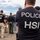 The image shows a group of law enforcement officers from HSI (Homeland Security Investigations), a division of U.S. Immigration and Customs Enforcement (ICE). Multiple agents are visible wearing dark navy t-shirts with 'POLICE HSI' printed on the back. The agents appear to be assembled outdoors, possibly during a briefing or staging for an operation.