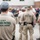 The image shows multiple U.S. Customs and Border Protection (CBP) Border Patrol Federal Agents gathered in an outdoor area, likely during a briefing or assembly. The foreground agent is wearing a tan uniform shirt with 'CBP BORDER PATROL FEDERAL AGENT' printed in green on the back. A large group of personnel, both in uniform and civilian clothing, are visible in the background.