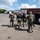 Several tactical police officers in full gear are walking across an industrial parking lot toward a large facility. The officers are wearing body armor, tactical vests labeled 'POLICE,' and carrying equipment, suggesting an active law enforcement operation. The scene takes place in a commercial/industrial area with trucks and a brick warehouse visible in the background.