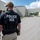 An ICE (Immigration and Customs Enforcement) officer is seen from behind, wearing a tactical vest marked 'POLICE ICE', standing in what appears to be an industrial or warehouse complex. The officer is equipped with standard law enforcement gear and appears to be observing a scene further down the facility's access road. Several other individuals are visible in the background near the building entrance.
