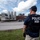 A law enforcement officer wearing a 'POLICE HSI' (Homeland Security Investigations) tactical vest and navy cap stands with his back to the camera, surveying an industrial facility. The officer appears to be monitoring or conducting surveillance of what looks like a food processing or manufacturing plant with large stainless steel tanks and industrial equipment. The scene suggests an active law enforcement operation at an industrial site.