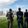 Three law enforcement officers are standing on a grassy area facing an industrial facility. Two agencies are visibly represented: CBP Border Patrol and HSI (Homeland Security Investigations). The officers appear to be conducting surveillance or an operational assessment of the facility.