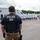 An HSI (Homeland Security Investigations) Police officer in tactical gear stands watch over a large group of individuals wearing white lab coats and hard hats in an outdoor industrial area. The scene appears to depict a law enforcement operation, likely a worksite raid or enforcement action. A Swift refrigerated trucking trailer is visible in the background.
