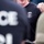 The image shows law enforcement personnel in a group setting. The central figure wears a black t-shirt bearing an HSI (Homeland Security Investigations) Special Agent badge insignia. An American flag patch is visible on the right sleeve of another individual in the background.