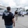 The image shows a law enforcement operation conducted by HSI (Homeland Security Investigations), a division of ICE. Multiple HSI/Police agents in tactical vests are overseeing a large group of workers wearing white lab coats and hard hats in an industrial or food processing facility parking area. This appears to be a worksite enforcement action at what looks like a processing plant.