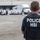 An HSI (Homeland Security Investigations) Police officer is seen from behind, observing a large group of individuals wearing white uniforms or protective clothing gathered in an industrial or warehouse area. A white semi-truck and loading dock are visible in the background. The scene appears to depict a law enforcement operation at a commercial or industrial facility.