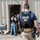 The image shows multiple law enforcement officers from HSI (Homeland Security Investigations) conducting what appears to be a field operation or raid at a metal-sided building marked 'S-1'. Officers are wearing tactical vests labeled 'POLICE HSI' and are entering or positioned near an open doorway. The scene depicts an active law enforcement operation in an outdoor industrial or commercial setting.