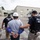 Two law enforcement officers wearing 'POLICE HSI' (Homeland Security Investigations) tactical vests are escorting a handcuffed individual wearing a white hard hat and gray t-shirt across an industrial facility parking lot. The scene appears to depict an arrest or detention operation at what looks like a warehouse or manufacturing facility. Additional personnel and vehicles are visible in the background.