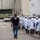 An HSI (Homeland Security Investigations) police officer is escorting a large group of workers wearing white protective gear, hard hats, and face shields along an outdoor corridor between industrial buildings. The workers appear to be food processing or industrial plant employees based on their protective equipment including white coats, rubber boots, and gloves. Additional law enforcement personnel are visible in the background near parked vehicles.