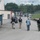 The image shows several individuals walking away from the camera along a paved road at what appears to be an industrial facility. Two officers wearing black 'POLICE' tactical vests are accompanied by civilians, including workers wearing hard hats and high-visibility clothing. The setting includes industrial buildings, large tanks, trucks, and a security fence in the background.