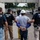The image shows HSI (Homeland Security Investigations) Police agents escorting a handcuffed individual wearing a white hard hat and work attire through what appears to be an industrial facility. Multiple agents in tactical vests marked 'POLICE HSI' are visible, along with a larger group of people in the background. This appears to document a law enforcement operation at a worksite.