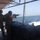 Two individuals in military-style attire are positioned on the stern deck of a moving vessel at sea. One person is actively firing a rifle while the other observes. Spent casings are visible in the air, indicating live fire is in progress.