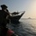 A heavily armed military or law enforcement personnel in tactical gear is positioned on an inflatable rigid hull boat (RHIB), observing or approaching a traditional wooden dhow vessel on open water. A larger naval vessel is visible in the background on the horizon. The scene appears consistent with a maritime interdiction or boarding operation.