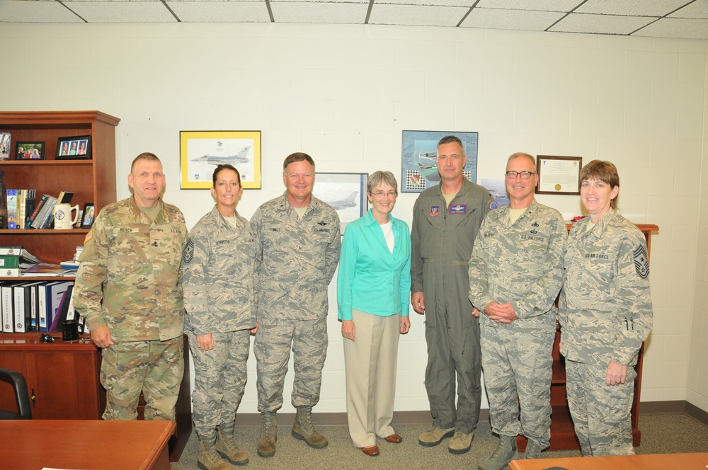 A group of seven individuals pose for a formal photo in what appears to be a military office setting. Six of the individuals are wearing military uniforms — a mix of Army combat uniforms and Air Force ABUs (Airman Battle Uniforms), along with one person in a flight suit — while one civilian woman in a teal blouse and khaki pants stands in the center. The setting suggests an official visit or meeting at a military installation.